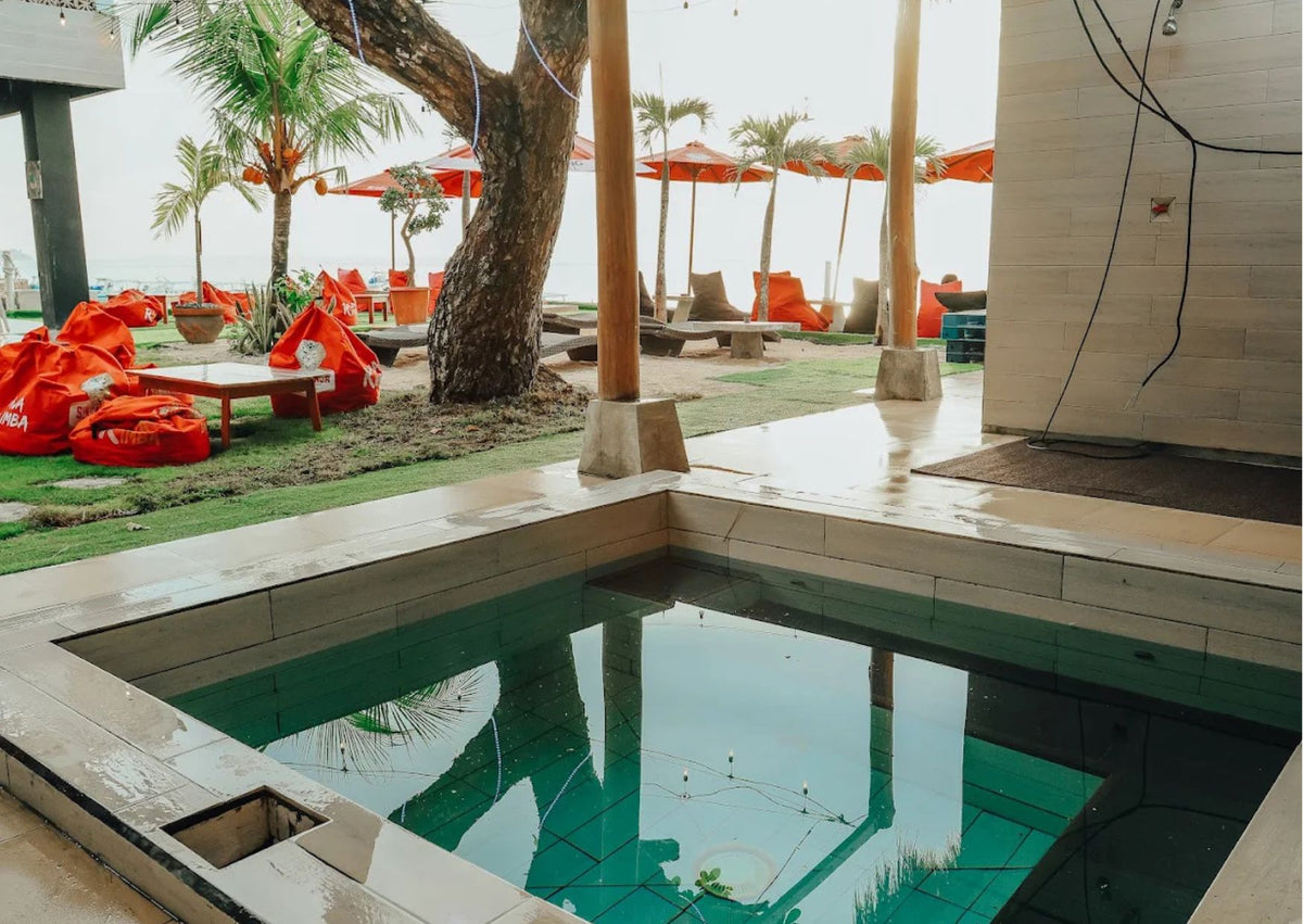 A rectangular outdoor ice bath with clear turquoise water, surrounded by light beige stone tiles. The pool is situated on a resort patio with palm trees, lounge chairs, and bright orange umbrellas in the background.