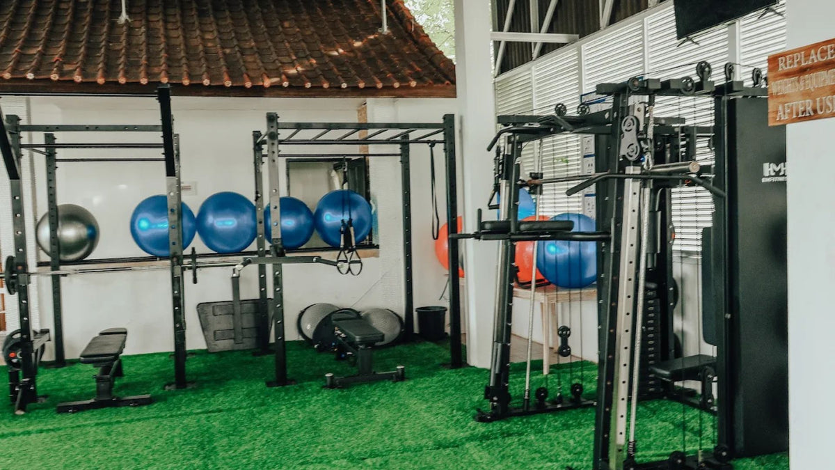 The gym interior of 3 Monkeys Fitness Centre in Lembongan, with multiple pieces of exercise equipment, including a pull-up bar rack, a cable machine, a bench, and several blue exercise balls mounted against a white wall under a tiled roof.