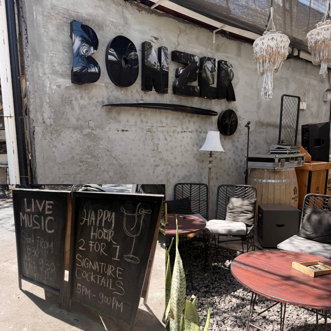 Outdoor patio seating area of a venue with a large black 'BONZER' sign on a concrete wall, black chalkboard signs advertising live music and happy hour in Jungut Batu, metal chairs, round tables, hanging lanterns, and stacked equipment on a gravel floor.