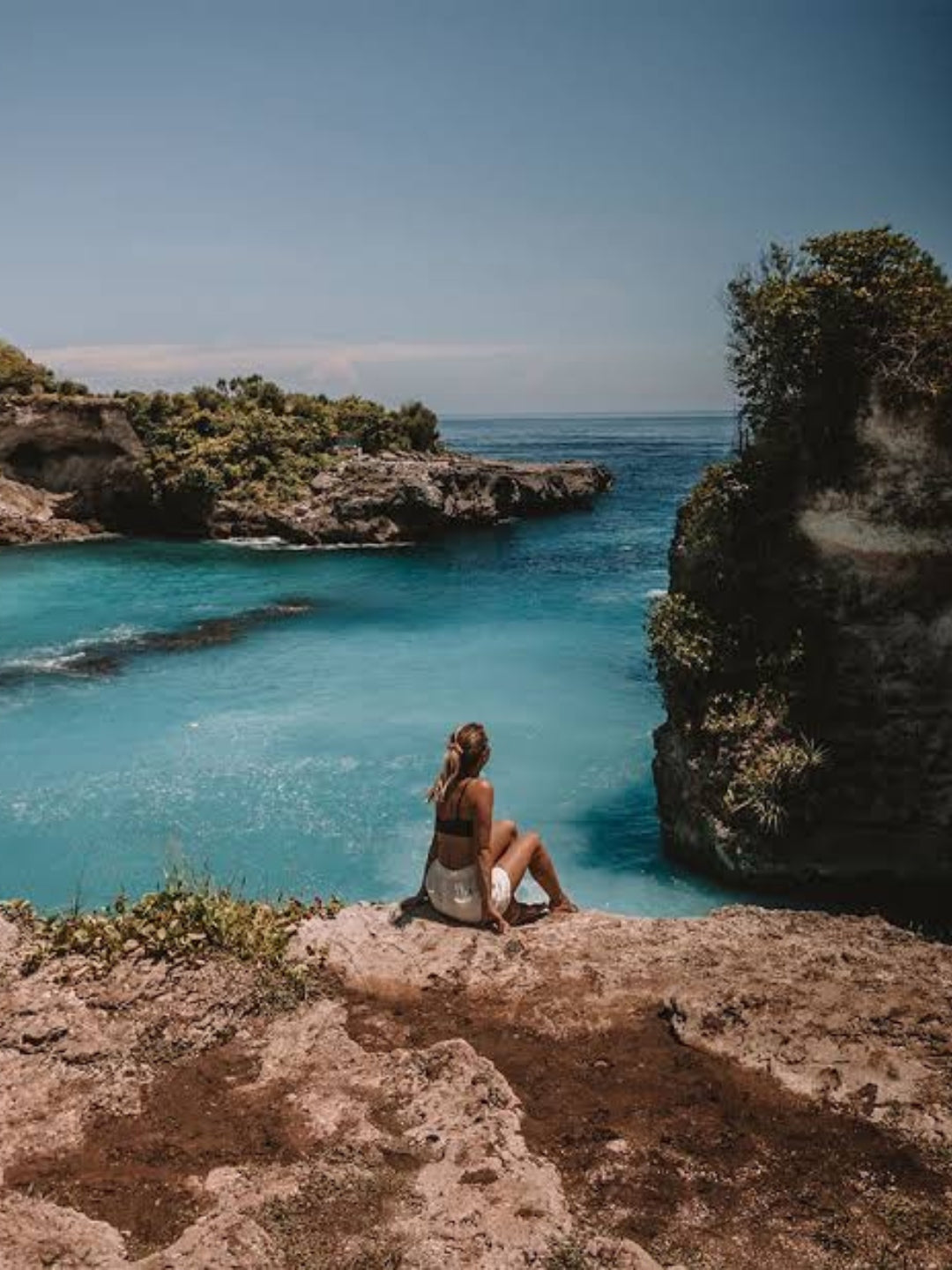A woman sits on a rocky cliff overlooking a turquoise ocean bay with rugged coastline and green vegetation under a clear blue sky.