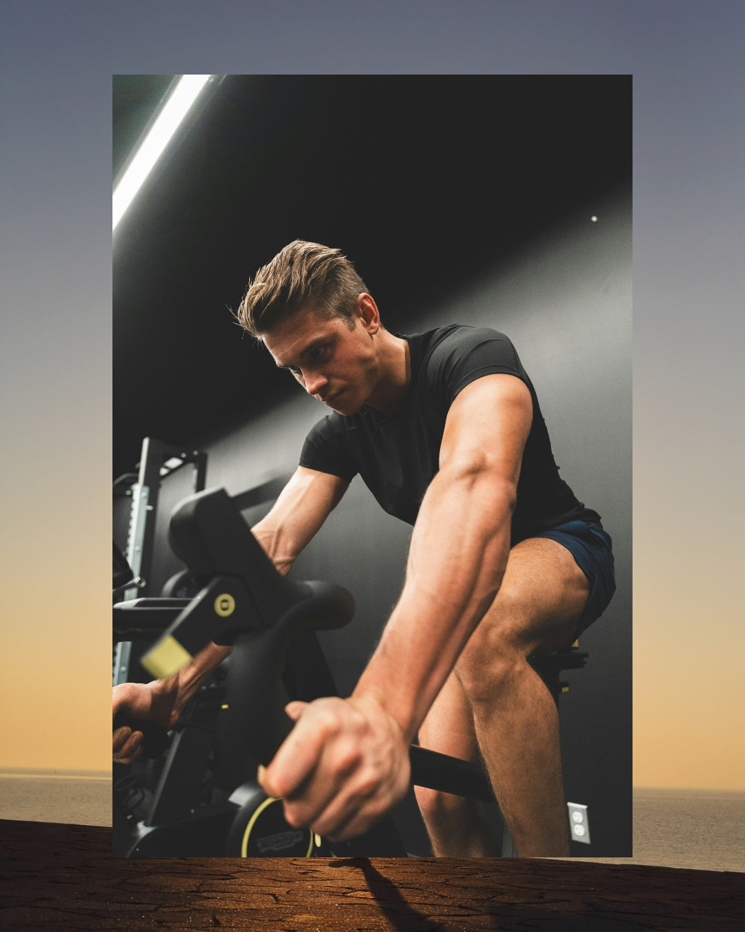 A man in a black athletic shirt and shorts riding a stationary exercise bike in a gym setting, shown within a vertical frame overlay on a sunset background.