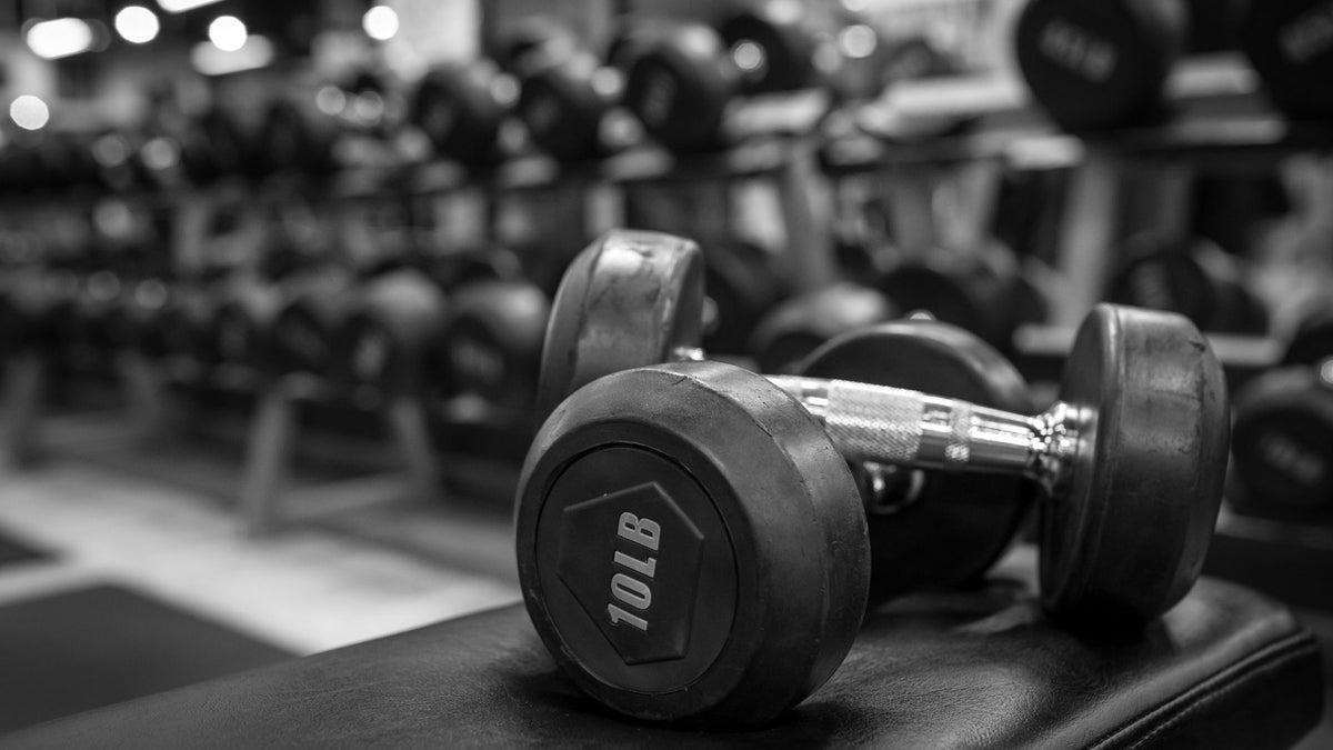 A 10LB dumbbell in the foreground on a gym bench, with rows of other dumbbells blurred in the background. The photo is in black and white, emphasizing the dark rubber weights and metallic handles.