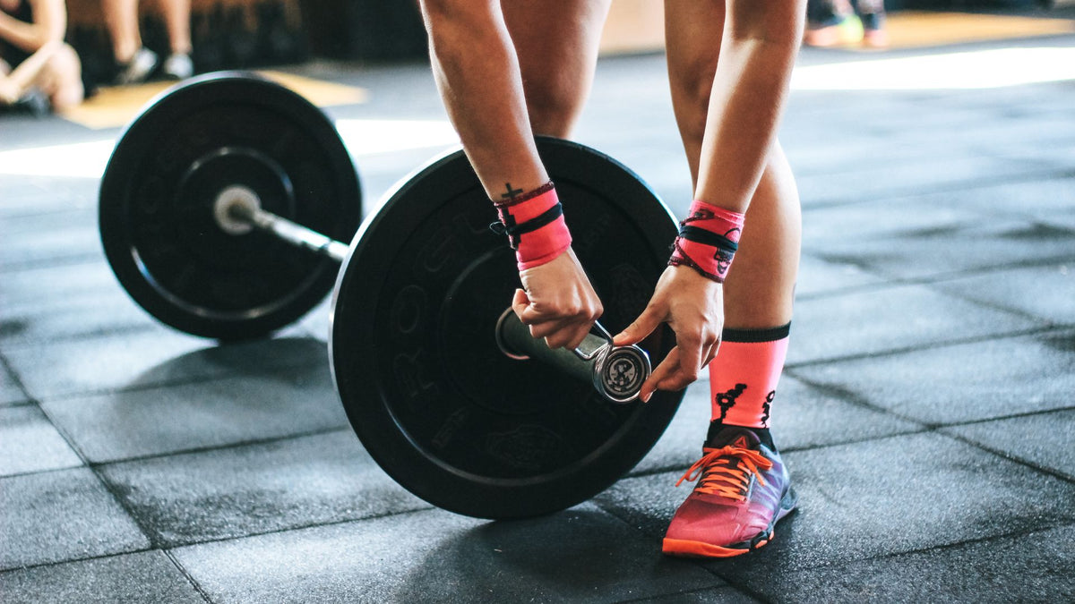 A close-up lifestyle shot of a person in a gym lifting a barbell, showing their hands, legs, and feet. The person is wearing pink and black wrist wraps, pink athletic socks, and pink and orange running shoes on dark rubber gym flooring.