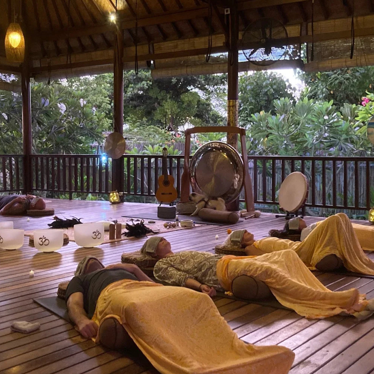 Several people lie on yoga mats on a wooden deck inside a pavilion, wrapped in yellow blankets, with yoga props, a large drum, and lush greenery visible in the background.
