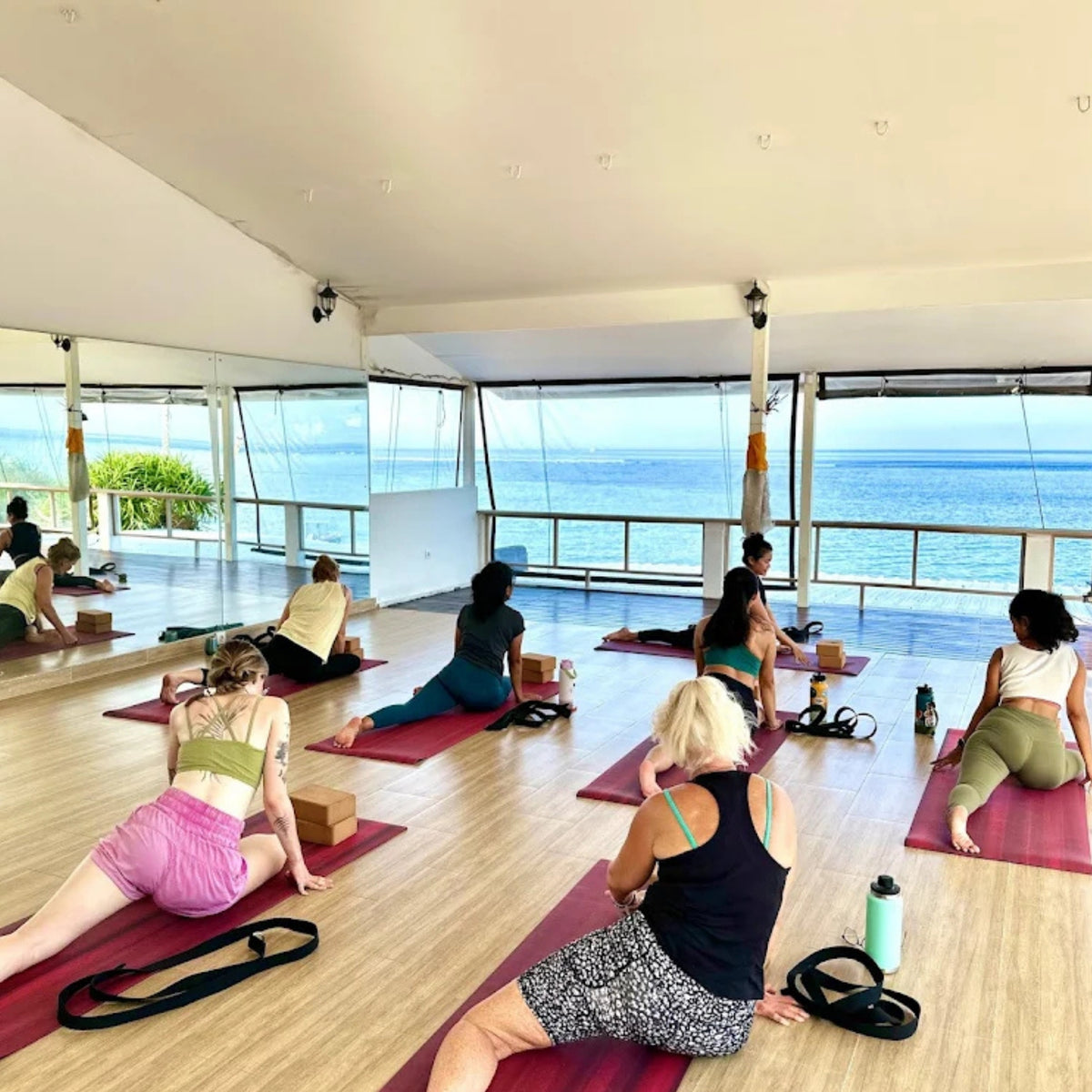 A group of people practicing yoga on pink mats in a bright studio with large windows overlooking the ocean.