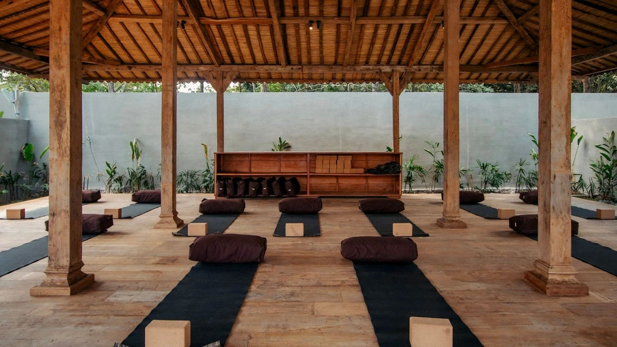 An open-air wooden pavilion with rows of cushioned meditation or yoga mats arranged on a stone floor, with a wooden bench and plants against a light gray wall in the background.