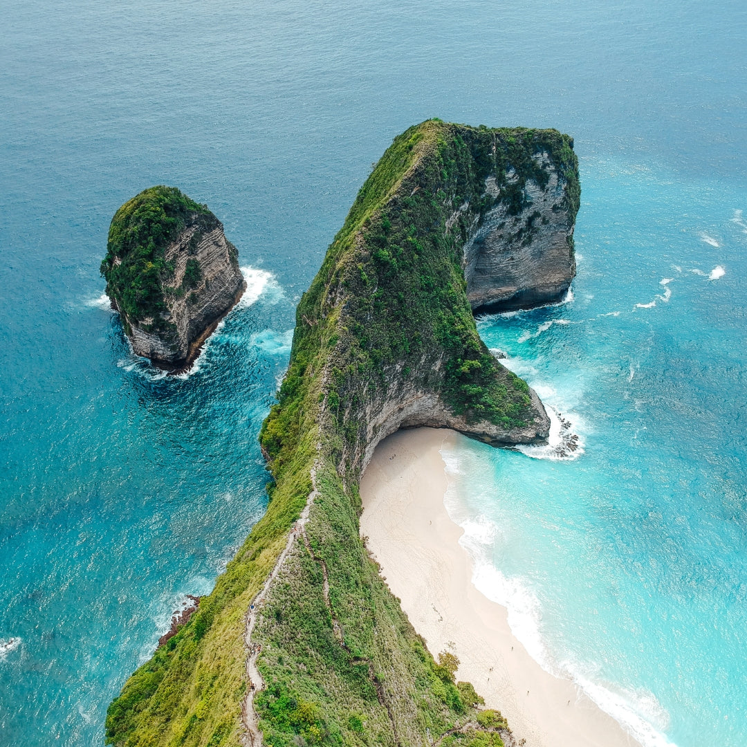 Aerial view of a dramatic T-Rex coastal rock formation with a narrow sandy beach, surrounded by turquoise and deep blue ocean water at Kelingking Beach in Nusa Pendia
