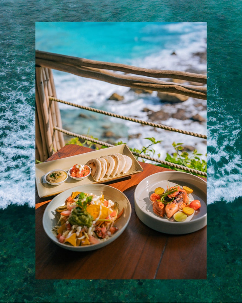 Two bowls of seafood and salad on a wooden table, with sliced fish on a tray in the background, set against an ocean view with a rustic wooden railing. The photo is framed by a teal ocean wave border, this is food served at Klyf Club on Nusa Ceningan.