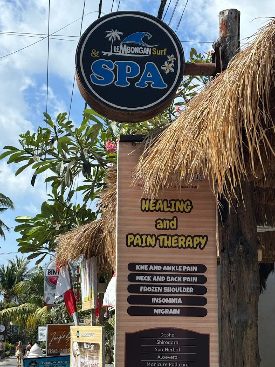 Outdoor sign for Lembongan Surf & Spa with a round black-and-white logo above a tall wooden panel reading 'Healing and Pain Therapy' and listing treatment areas such as knee and ankle pain, frozen shoulder, and migraines. A thatched roof and tropical trees are visible in the background.