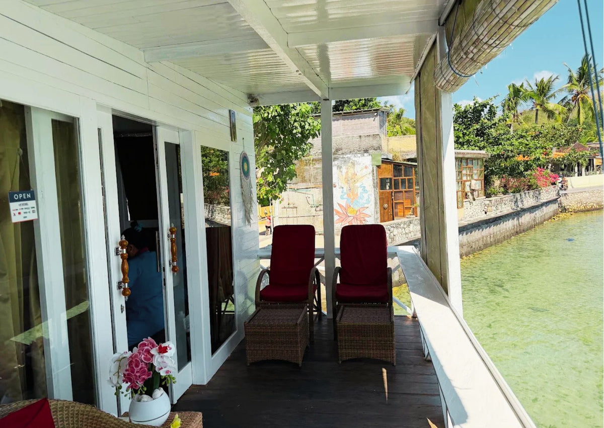A covered outdoor porch with two red cushioned lounge chairs and wicker ottomans facing a canal view. The porch has white walls and glass doors, with tropical greenery and water visible outside. The balcony of Leony Ocean Spa on Nusa Ceningan
