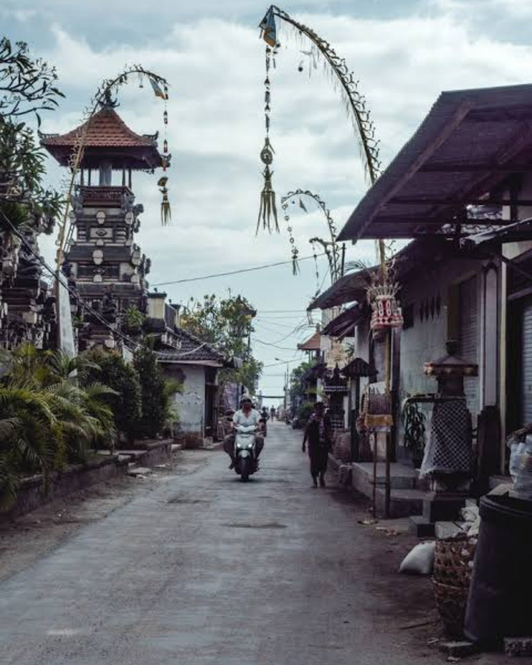 A narrow street in a traditional Balinese village with a tall pagoda, hanging arched ceremonial banners, a motorbike rider, and pedestrians under a cloudy sky in Lembongan Village.