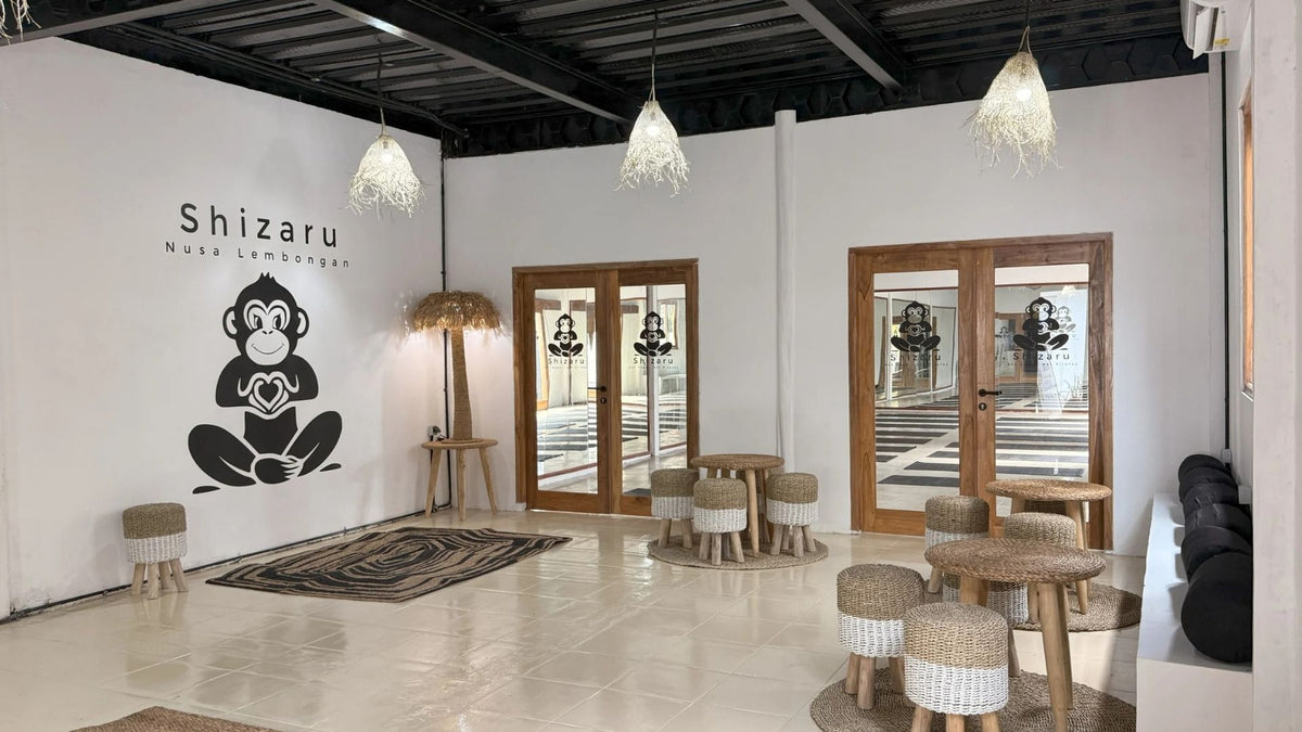 A spacious, modern indoor lounge or studio space with white walls, black ceiling beams, and hanging pendant lights. The left wall features a large black graphic of a meditating monkey with the text 'Shizaru Nusa Lembongan'.