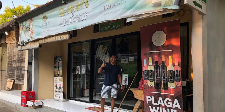 Man standing outside of Q-Mart wine shop with promotional posters and products displayed. Located Jungut Batu