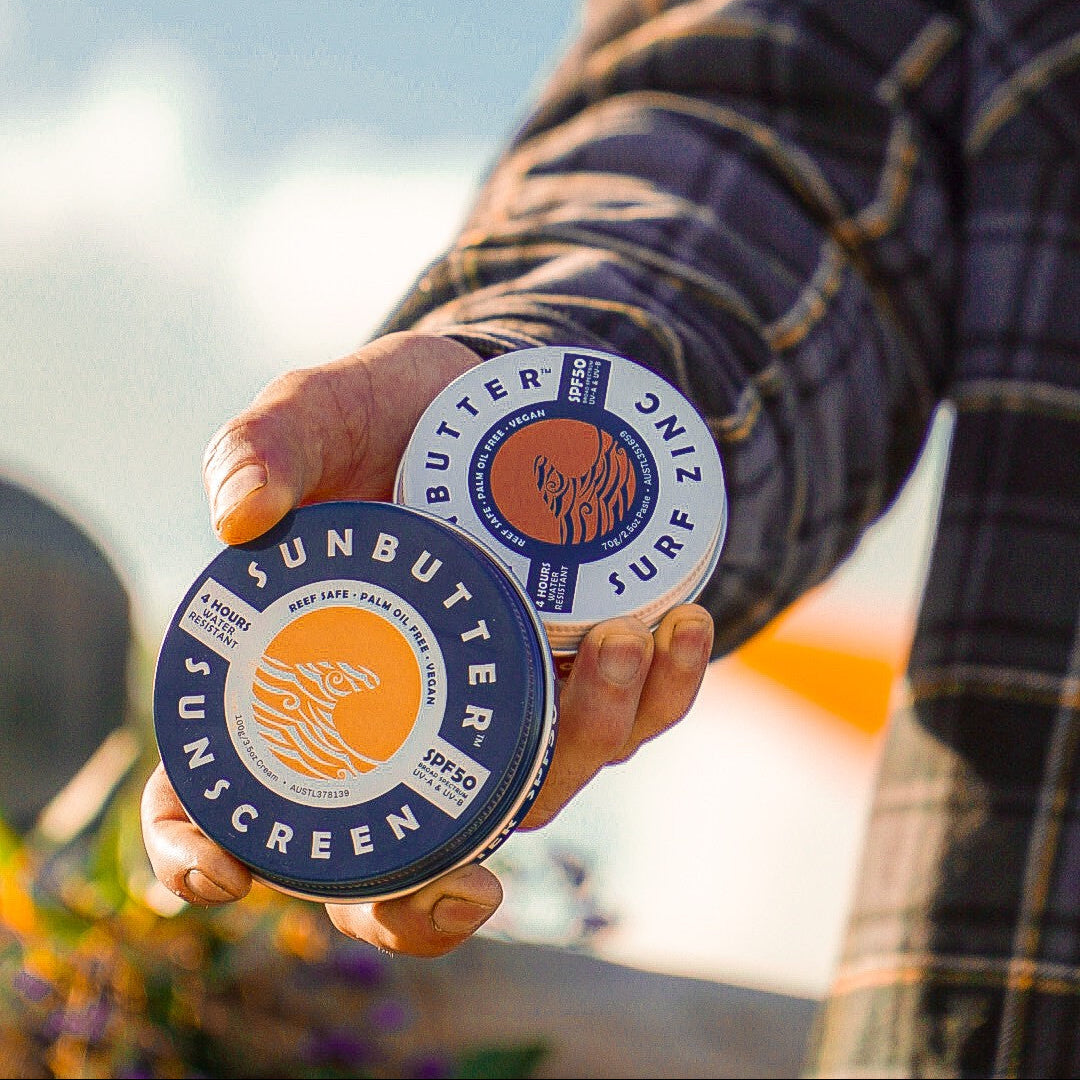 A person outdoors holds two small round tins of sunscreen butter. One tin is labeled 'SUNBUTTER SUNSCREEN' and the other 'BUTTER SURF'.