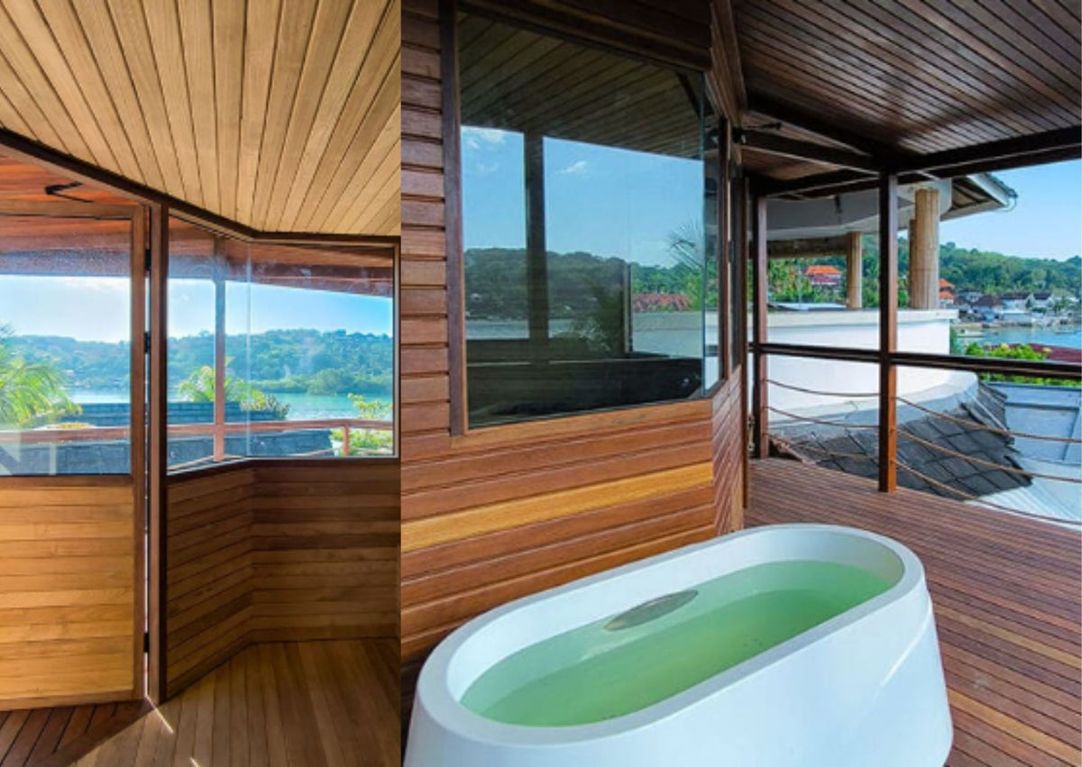 A white freestanding oval bathtub filled with greenish water, placed on a wooden deck inside a modern open-air pavilion with large windows overlooking water and hills. The Segara Wellness Centre.
