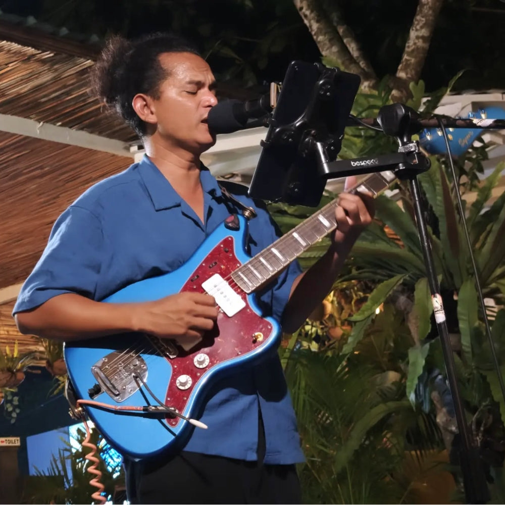 A man playing a blue electric guitar with a red pickguard, singing into a microphone in an outdoor nighttime setting with tropical plants in the background at Agus Shipwrecks Beach Bar, Nusa Lembongan
