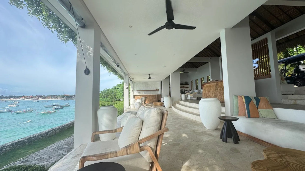 The modern outdoor patio with lounge chairs, at Spring Spa, Lembongan, with a built-in sofa, and a large window overlooking a tropical ocean with boats. The space features white walls, a tiled floor, a ceiling fan, and a view deck leading to an interior.