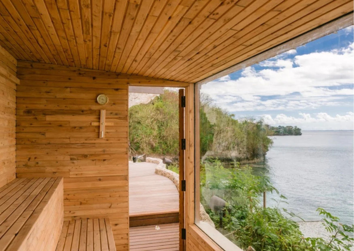 A wooden sauna interior with a large glass window overlooking a scenic coastal landscape with water, greenery, and sky from the Sun Deck at Lago