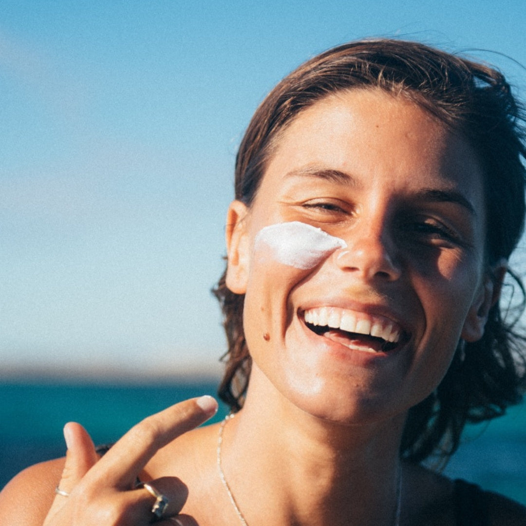 A close-up lifestyle shot of a woman outdoors by the sea, smiling broadly with white sunscreen cream applied to her cheek. She is wearing a delicate silver chain necklace and a ring, with her hand raised near her face.