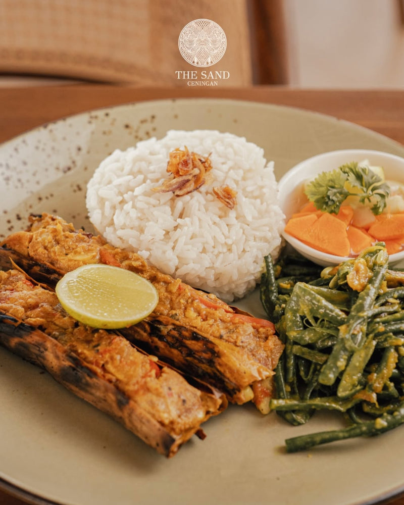 A plated meal featuring grilled fish with a lime wedge, a mound of white rice, stir-fried green beans, and sliced orange vegetables, served on a beige speckled plate. Food served at The Sand resto in Nusa Ceningan