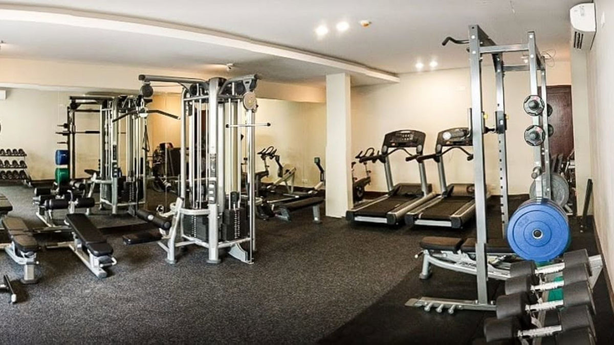 A spacious indoor gym with multiple exercise machines, including cable machines, treadmills, weight benches, and a barbell rack on a dark carpeted floor.