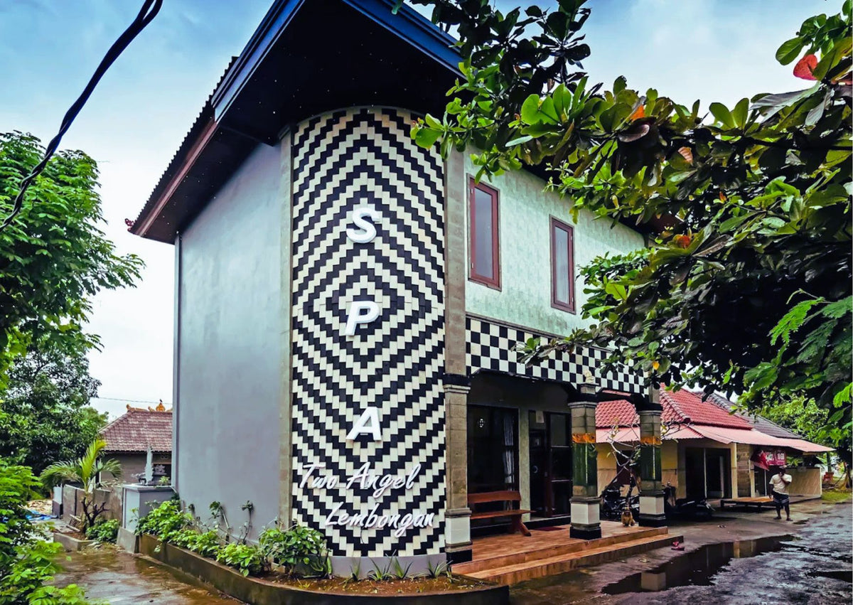 A two-story building with a bold black-and-white checkered facade and large white 'Two Angel Lembongan SPA' lettering on the side, photographed outdoors with trees, sky, and a wet street in view.