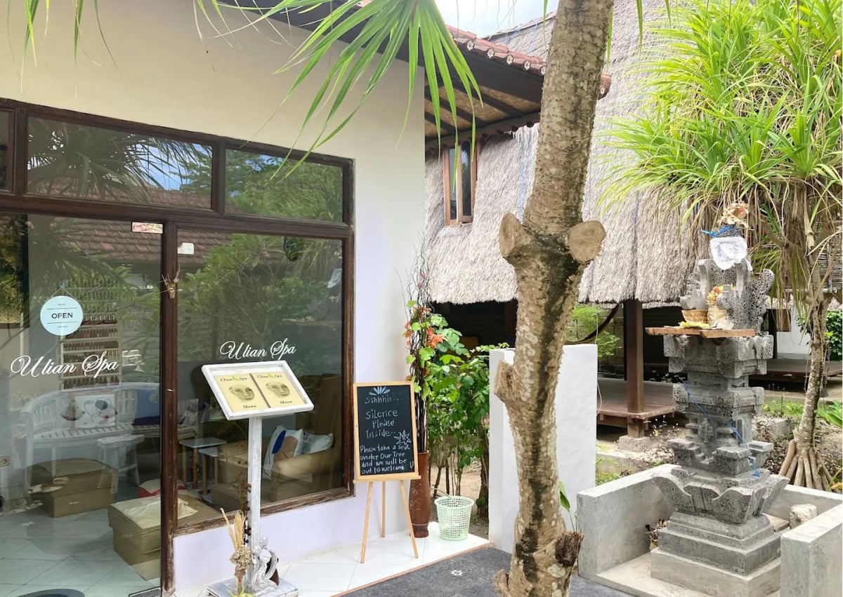 A tropical outdoor view of a Ulian Spa  wellness centre storefront with large glass doors, a thatched-roof building in the background, palm leaves overhead, and a stone statue on the right.