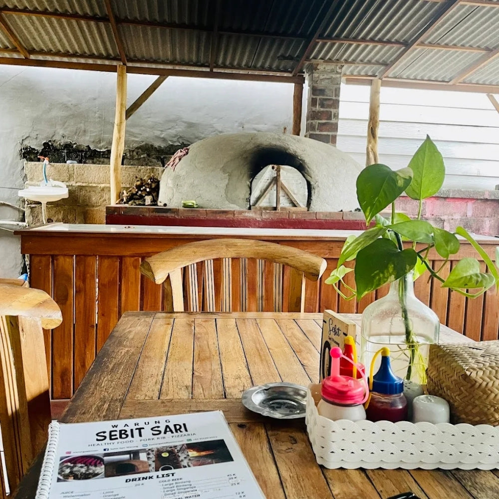 A rustic wooden dining table in an outdoor covered patio setting, with a menu titled 'SEBIT SARI' in the foreground, a potted green plant in a glass vase, and a white tray holding condiments and a woven basket. In the background, a large clay oven is visible under a corrugated metal roof.