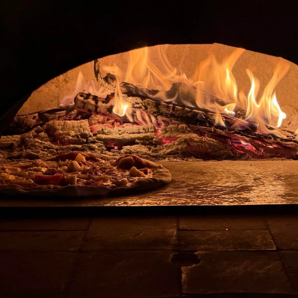 The pizza baking scene inside a wood-fired oven, with glowing orange flames and logs behind a pizza resting on the oven floor. Wood Fired pizza at Warung Sebit Sari