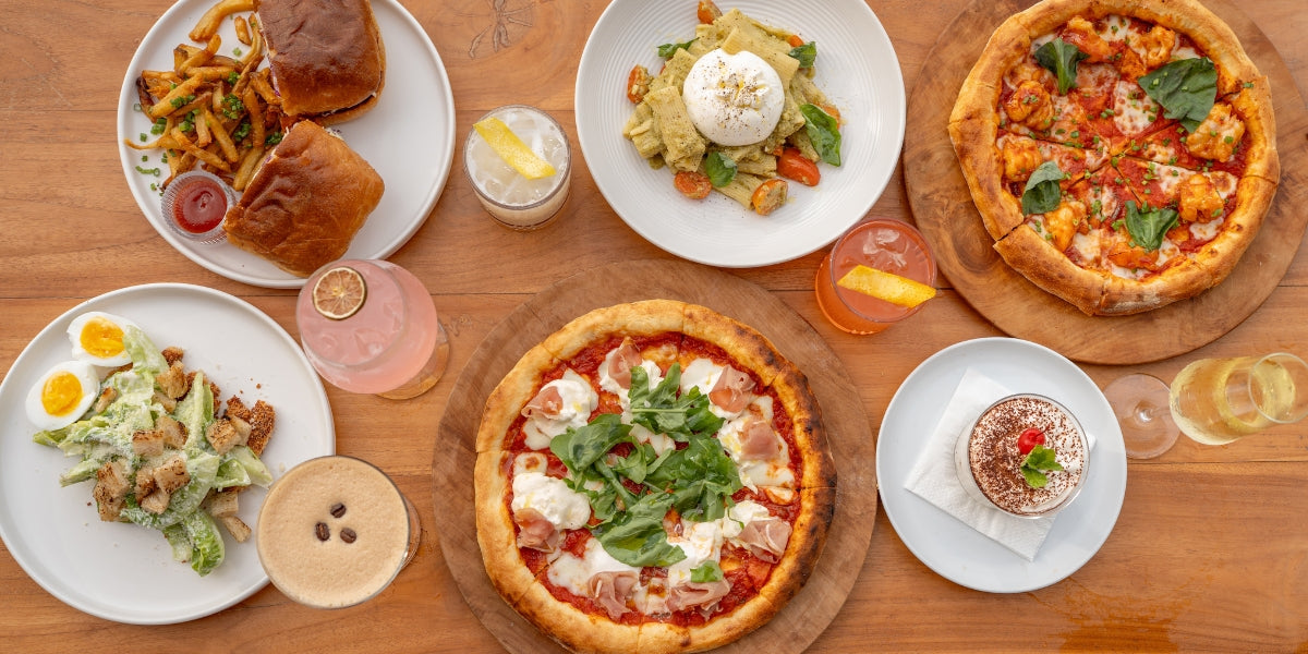 Assorted dishes including pizzas, salads, and pastries on a wooden table being served at Karana Pool Bar and Restaurant