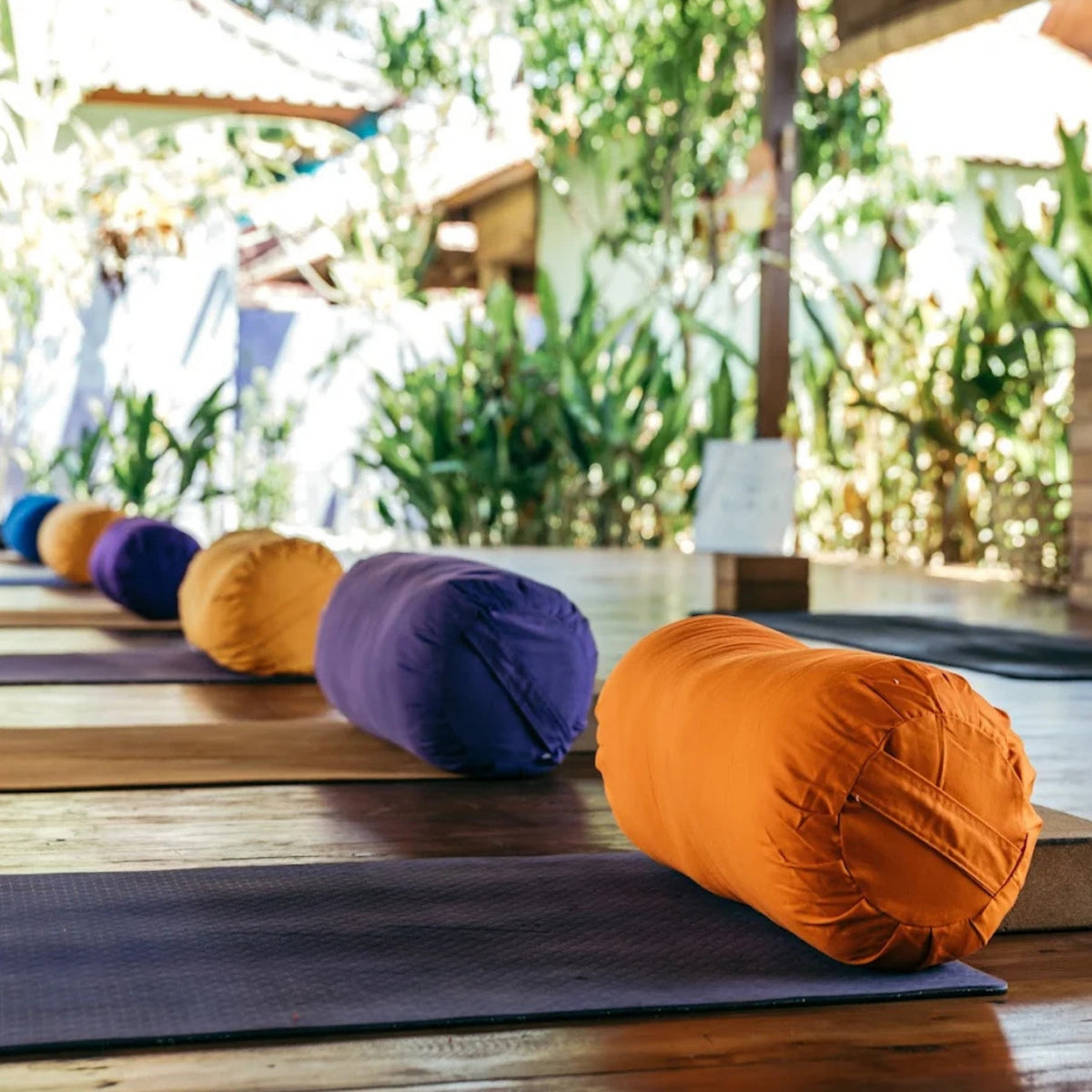 A row of colorful cylindrical yoga support pillows in orange, purple, and mustard yellow arranged on dark yoga mats on a wooden deck with a blurred outdoor garden background.