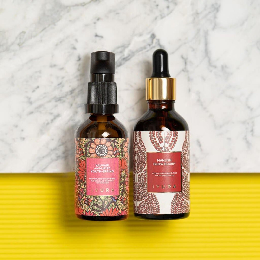 Two amber glass cosmetic dropper bottles from the brand ayurveda, displayed side-by-side on a bright yellow ribbed surface with a white marble background. The bottle on the left has a pink and orange floral patterned label and a black spray top.